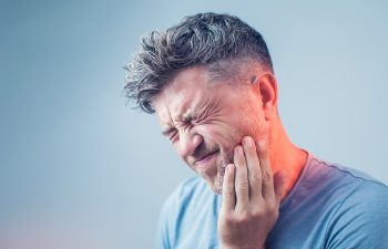 A man with short gray hair holds his cheek with a pained expression, indicating a toothache or facial discomfort.